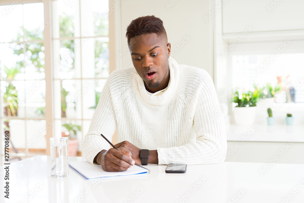 African american student man writing on a paper using a pencil scared in shock with a surprise face, afraid and excited with fear expression