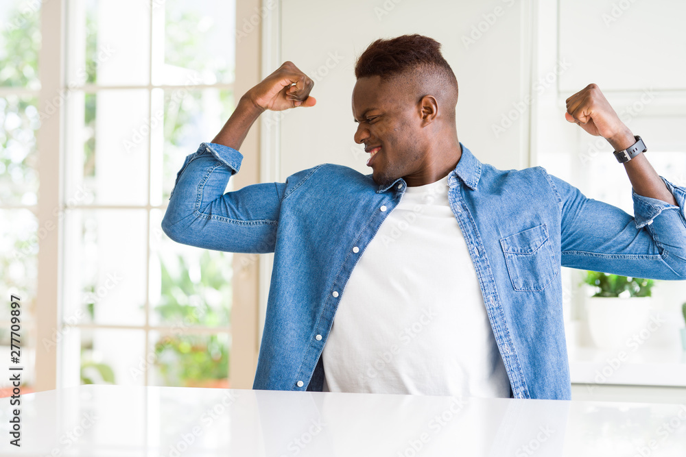 Fototapeta premium Handsome african american man at home showing arms muscles smiling proud. Fitness concept.