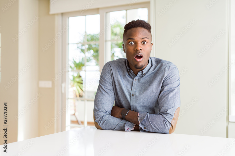 Handsome african american man on white table afraid and shocked with surprise expression, fear and excited face.