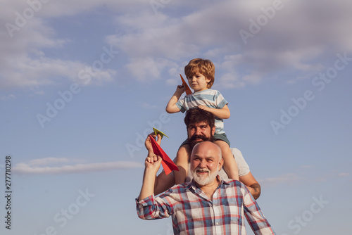 Happy grandfather father and grandson with toy paper airplane over blue sky and clouds background. Three men generation. Kids playing with simple paper planes on sunny day.
