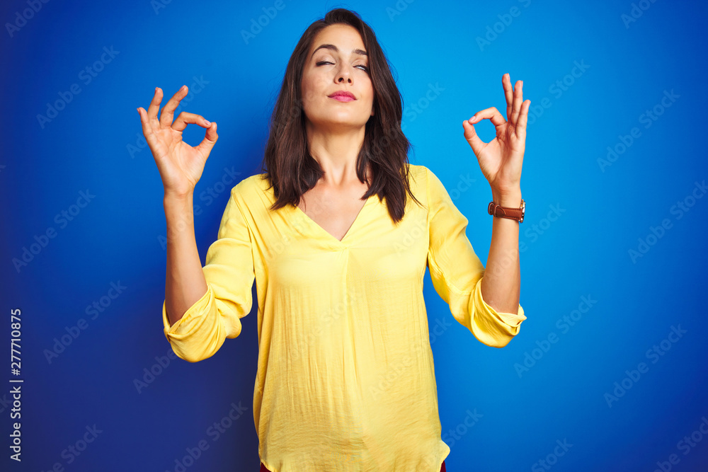 Young beautiful woman wearing yellow t-shirt standing over blue isolated background relax and smiling with eyes closed doing meditation gesture with fingers. Yoga concept. Young beautiful woman wearing yellow t-shirt standing over blue isolated background relax and smiling with eyes closed doing meditation gesture with fingers. Yoga concept.