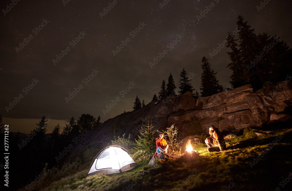 Night summer camping in the mountains. Happy couple hikers having a rest together, sitting beside campfire and glowing tourist tent. On background big boulder, forest and beautiful night starry sky. - obrazy, fototapety, plakaty 