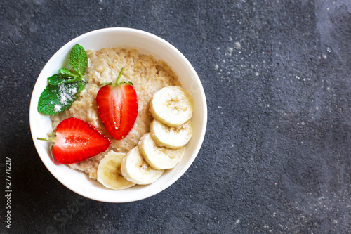 Fresh oatmeal with strawberries and banana slices in a white plate stands on a dark concrete background. On the right there is free space for inscriptions.