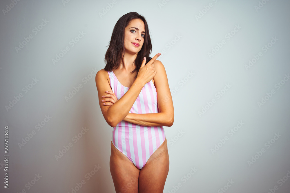 Young beautiful woman wearing striped pink swimsuit swimwear over isolated background Pointing with hand finger to the side showing advertisement, serious and calm face Young beautiful woman wearing striped pink swimsuit swimwear over isolated background Pointing with hand finger to the side showing advertisement, serious and calm face