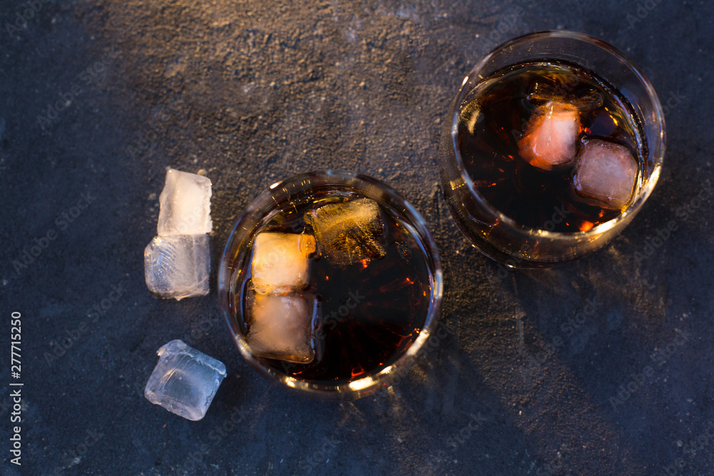 Two glasses with cocktails: rum cola and whiskey cola stand on a gray blue concrete textured background and there are a few ice cubes lying around Two glasses with cocktails: rum cola and whiskey cola stand on a gray blue concrete textured background and there are a few ice cubes lying around