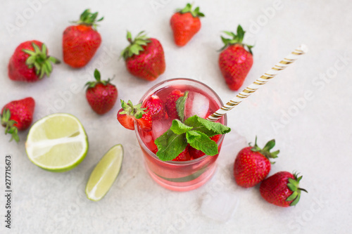 On a light background there is a strawberry cocktail in a glass with ice and mint, a bowl with strawberries and lime slices