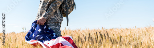 Panoramic shot of patriotic soldier in military uniform holding American flag while standing in field