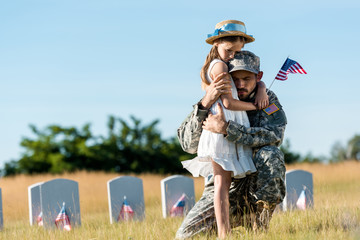 military man with closed eyes hugging daughter near headstones in graveyard