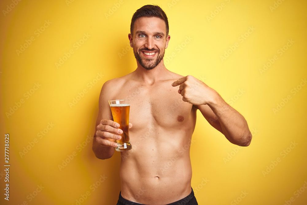 Young handsome shirtless man drinking a pint of beer over isolated yellow background with surprise face pointing finger to himself