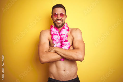 Young handsome shirtless man wearing sunglasses and pink hawaiian lei over yellow background happy face smiling with crossed arms looking at the camera. Positive person.