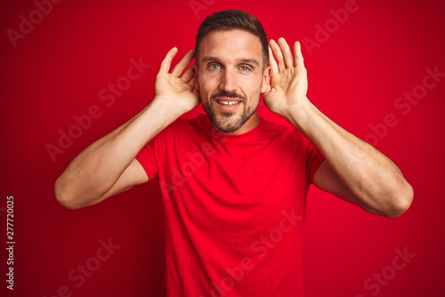 Young handsome man wearing casual t-shirt over red isolated background Trying to hear both hands on ear gesture, curious for gossip. Hearing problem, deaf