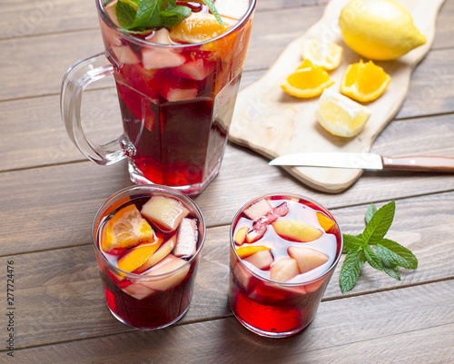 Two glasses and a pitcher with sangria and fruits next to table with lemon and orange cut on top next to knife. Sangria, a refreshing wine drink typical of Spain.