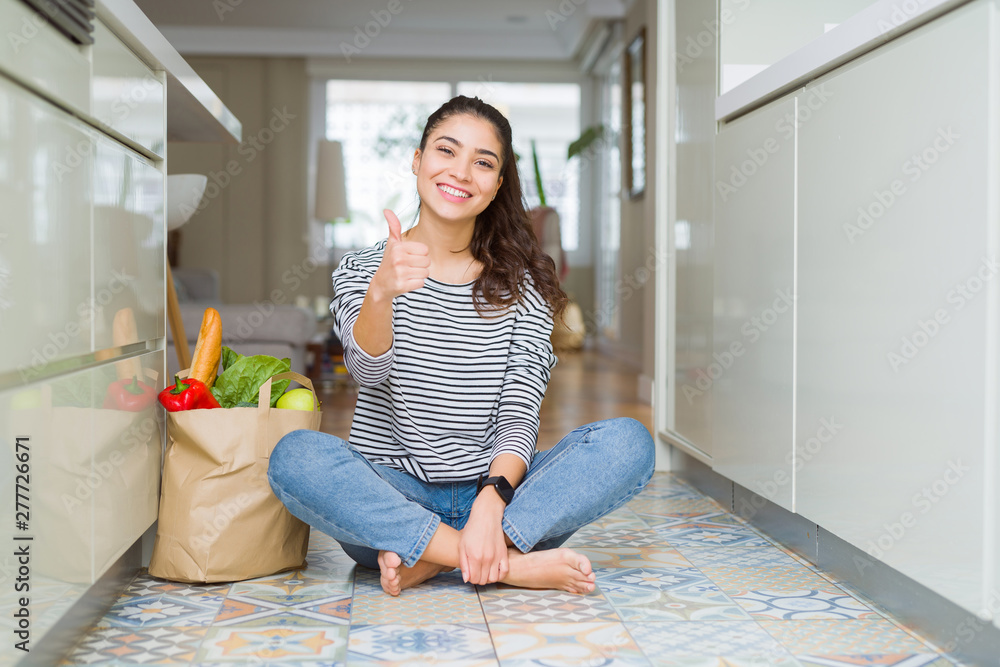 Young woman sitting on the kitchen floor with a paper bag full of fresh groceries doing happy thumbs up gesture with hand. Approving expression looking at the camera with showing success. Young woman sitting on the kitchen floor with a paper bag full of fresh groceries doing happy thumbs up gesture with hand. Approving expression looking at the camera with showing success.