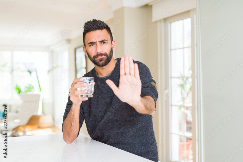 Handsome hispanic man drinking a fresh glass of water with open hand doing stop sign with serious and confident expression, defense gesture