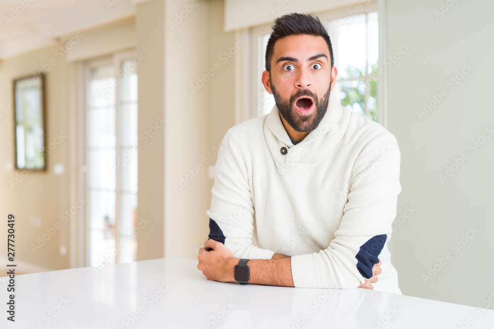 Handsome hispanic man wearing casual white sweater at home afraid and shocked with surprise expression, fear and excited face.