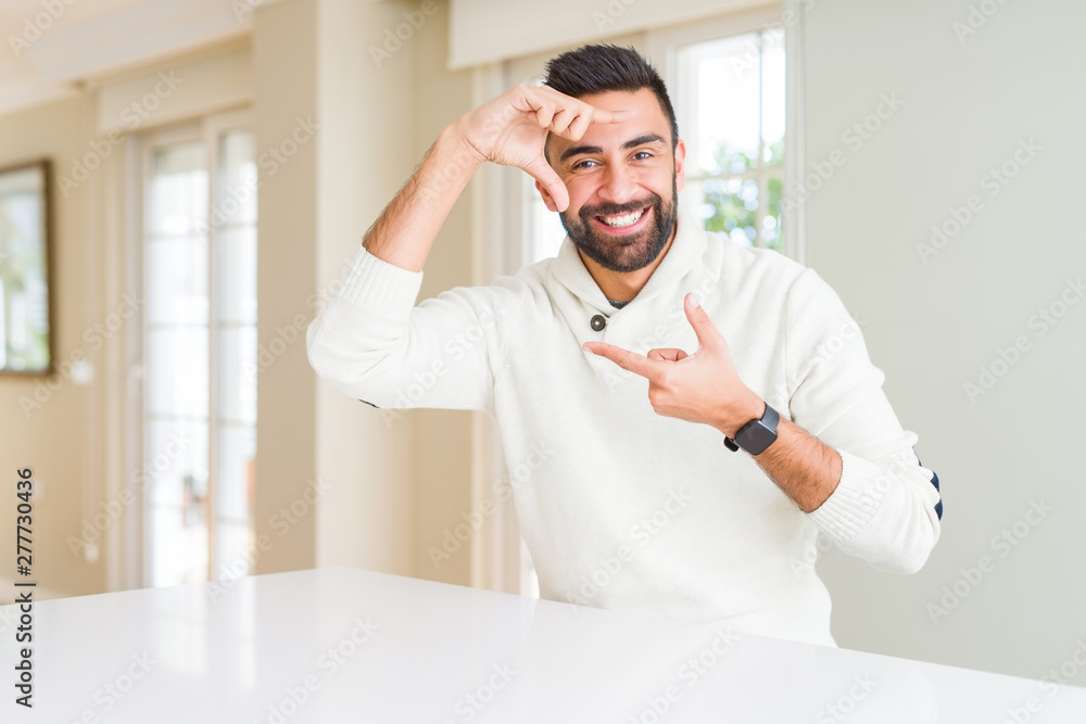 Fototapeta premium Handsome hispanic man wearing casual white sweater at home smiling making frame with hands and fingers with happy face. Creativity and photography concept.