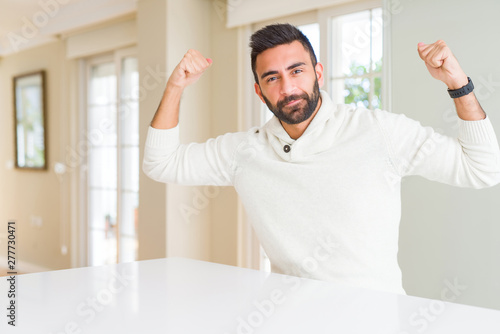 Handsome hispanic man wearing casual white sweater at home showing arms muscles smiling proud. Fitness concept.