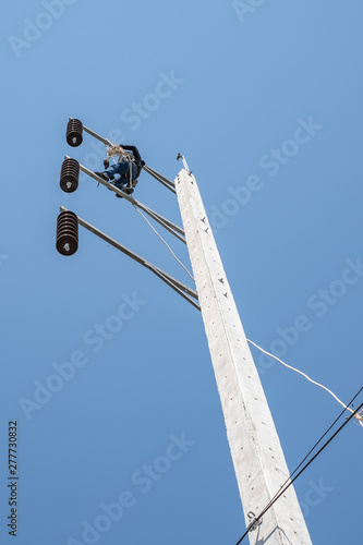 Electrician climbing very high electric power pole with safety belt to install new wire in very hot day with blue sky.