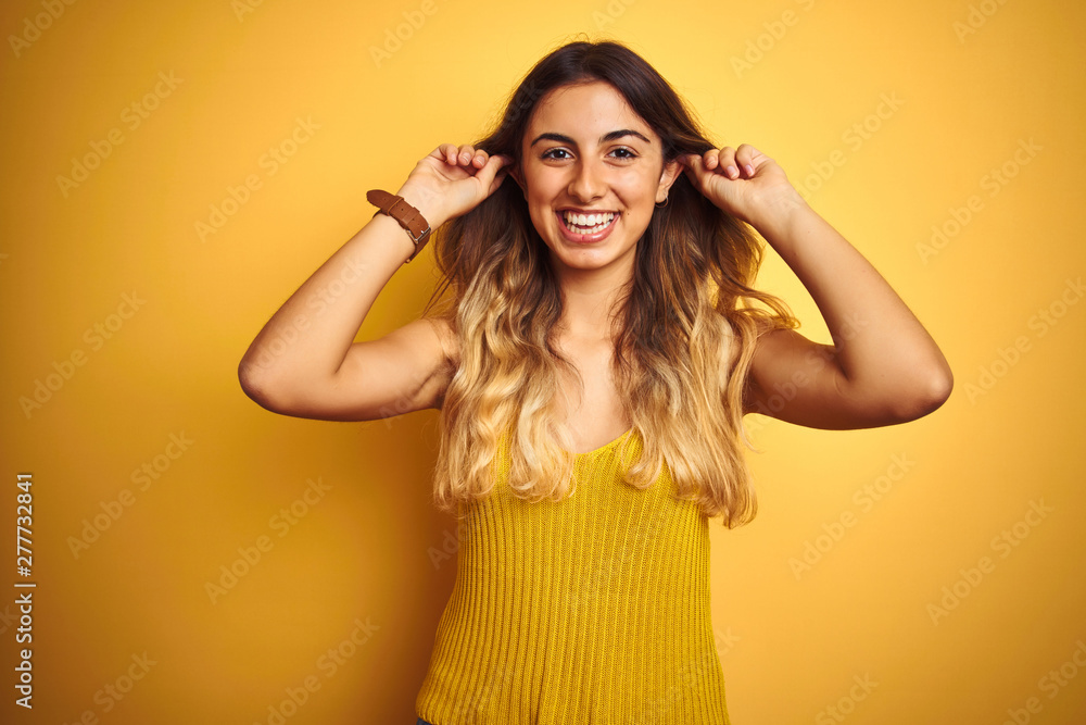 Young beautiful woman wearing t-shirt over yellow isolated background Smiling pulling ears with fingers, funny gesture. Audition problem Young beautiful woman wearing t-shirt over yellow isolated background Smiling pulling ears with fingers, funny gesture. Audition problem