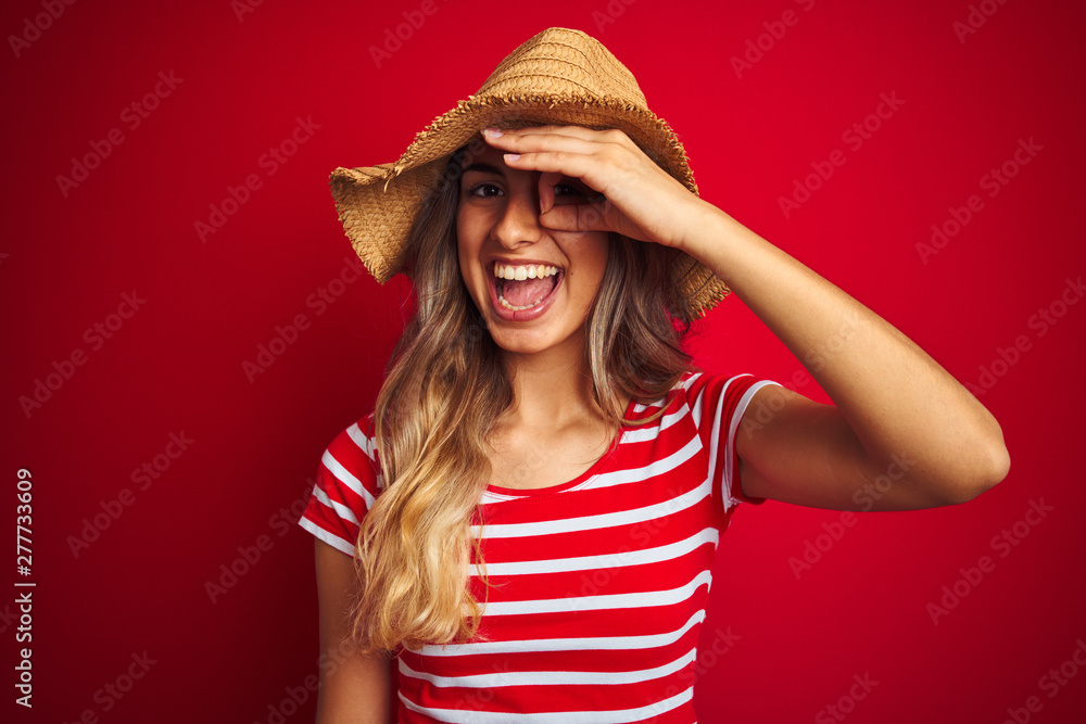 Young beautiful woman wearing summer hat over red isolated background with happy face smiling doing ok sign with hand on eye looking through fingers Young beautiful woman wearing summer hat over red isolated background with happy face smiling doing ok sign with hand on eye looking through fingers