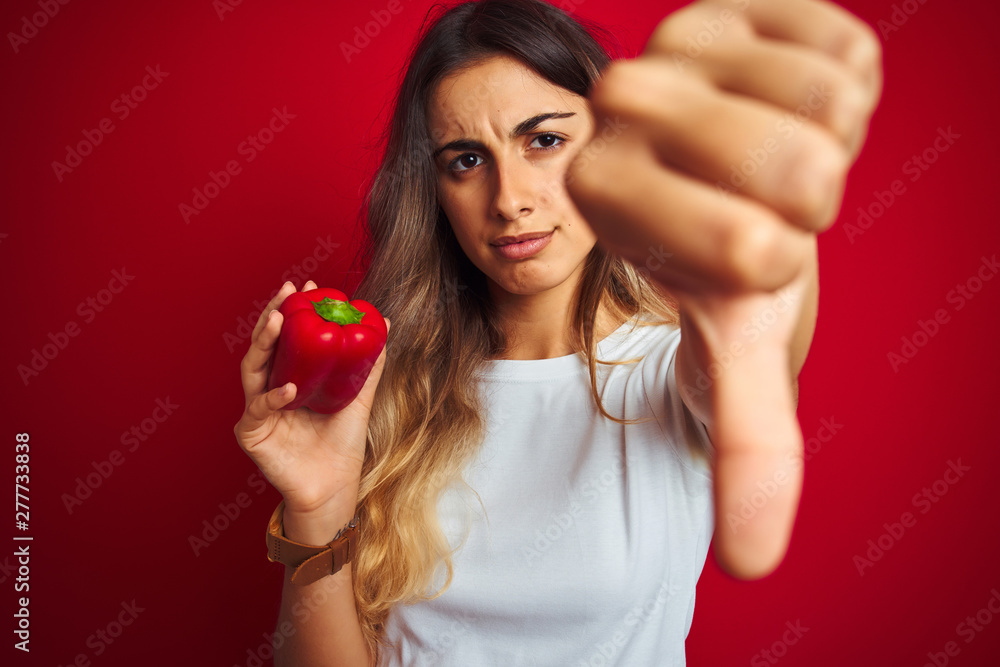 Young beautiful woman holding pepper over red isolated background with angry face, negative sign showing dislike with thumbs down, rejection concept Young beautiful woman holding pepper over red isolated background with angry face, negative sign showing dislike with thumbs down, rejection concept