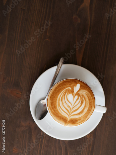 Overhead fresh brewed cappuccino coffee in white ceramic cup, saucer, spoon with beautiful handmade latte art on wooden table in cozy city cafe ready to drink