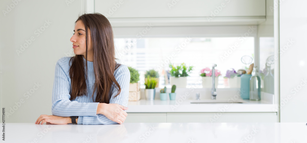 Wide angle picture of beautiful young woman sitting on white table at home looking to side, relax profile pose with natural face with confident smile. Wide angle picture of beautiful young woman sitting on white table at home looking to side, relax profile pose with natural face with confident smile.