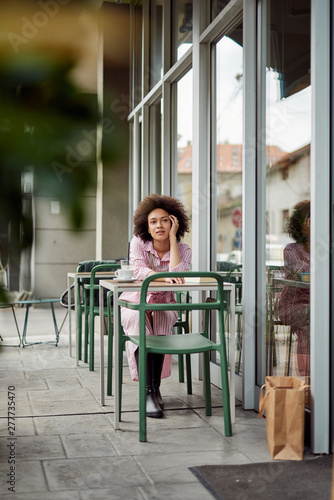 Beautiful mixed race woman with curly hair and dressed in pink striped dress leaning on table and thinking. Cafe exterior.