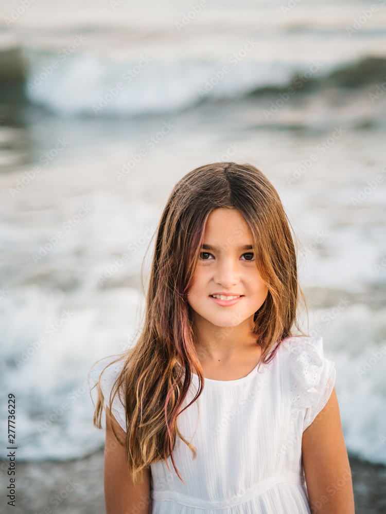 Portrait of charming little girl in white dress standing in water on sandy beach and looking at camera Portrait of charming little girl in white dress standing in water on sandy beach and looking at camera