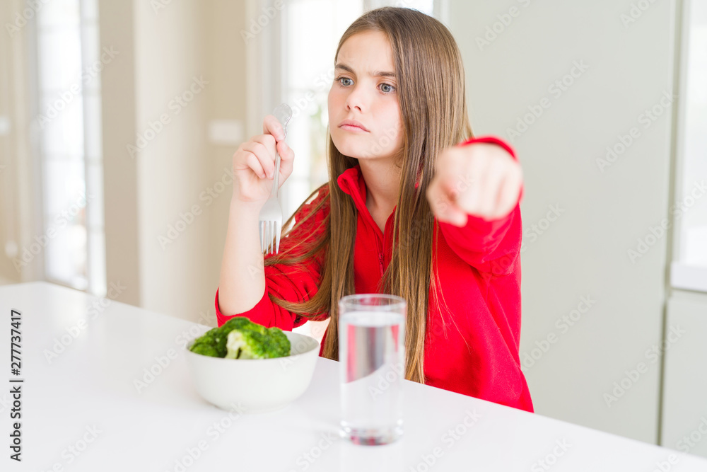 Beautiful young girl kid eating fresh broccoli and drinking water pointing with finger to the camera and to you, hand sign, positive and confident gesture from the front Beautiful young girl kid eating fresh broccoli and drinking water pointing with finger to the camera and to you, hand sign, positive and confident gesture from the front