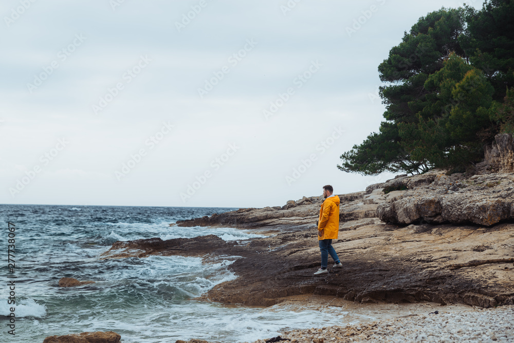 man walking by rocky beach in yellow raincoat. overcast windy weather