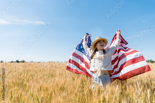 cheerful child in straw hat holding american flag in golden field