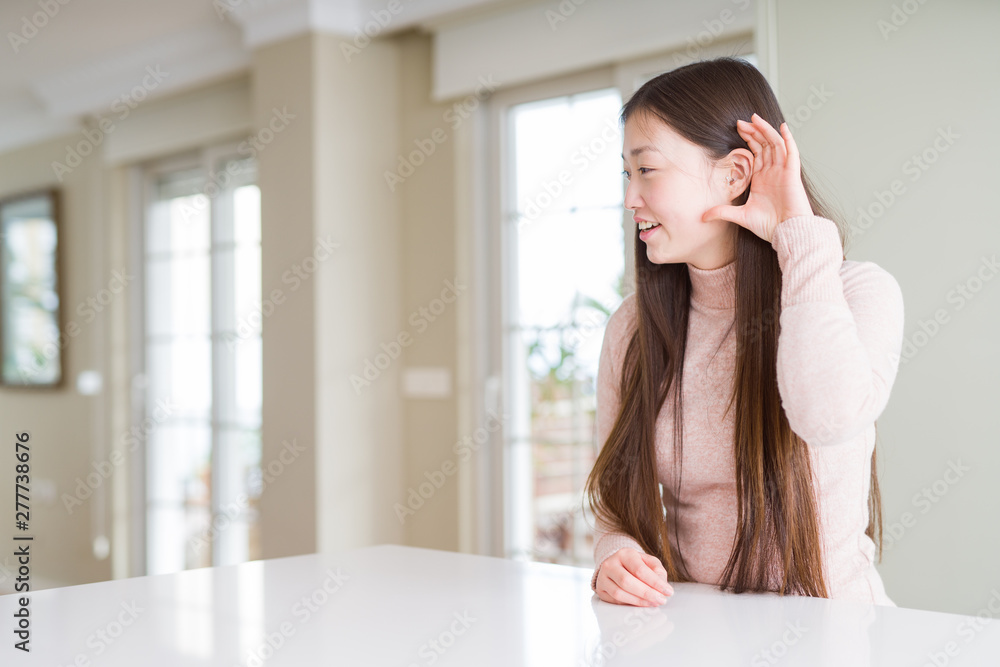 Beautiful Asian woman wearing casual sweater on white table smiling with hand over ear listening an hearing to rumor or gossip. Deafness concept. Beautiful Asian woman wearing casual sweater on white table smiling with hand over ear listening an hearing to rumor or gossip. Deafness concept.