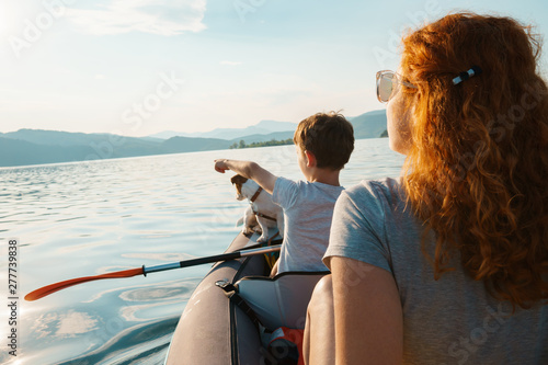 Happy young woman and her son with a dog Jack Russell Terrier are looking into the distance, rowing in an inflatable kayak on the water of mountain lake with a beautiful orange sunset. Family Weekend