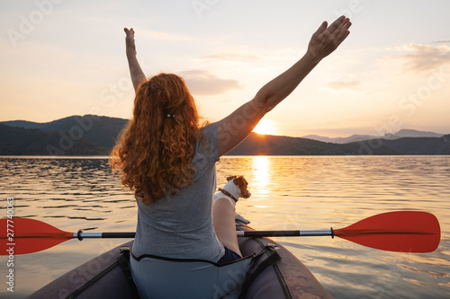 Happy young woman with her dog Jack Russell Terrier paddling on an inflatable kayak on the water of a large mountain lake against a beautiful orange sunset. Family Sports Weekend