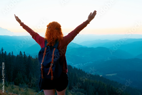 A relaxed red-haired woman in a dress is standing on top of a high mountain against the backdrop of a large mountain and spinning with happiness, raising her hands up. Bright sunset on the mountains