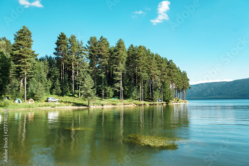 Panorama of a beautiful lake on a bright sunny day. Pine forest on the lake by the water. Clear clear water, blue sky in summer. Camping on the lake, tents and awnings. Tourism