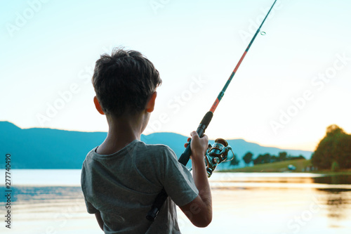 A happy boy is standing on the shore of a beautiful lake and fishing with a fishing rod against the background of a bright sunset in the summer. Child on summer vacation with family. Tourism. Camping