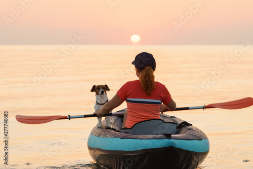 Red-haired young woman is rowing on an inflatable kayak by the sea with a dog Jack Russell Terrier on a background of pink sunrise in beautiful nature. Great disk of the rising sun. Sun rays. Sport