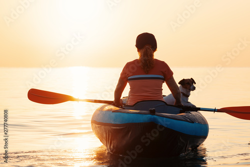 Red-haired young woman is rowing on an inflatable kayak by the sea with a dog Jack Russell Terrier on a background of pink sunrise in beautiful nature. Great disk of the rising sun. Sun rays. Sport