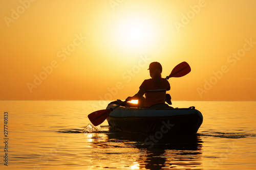 Red-haired young woman is rowing on an inflatable kayak by the sea with a dog Jack Russell Terrier on a background of pink sunrise in beautiful nature. Great disk of the rising sun. Sun rays. Sport