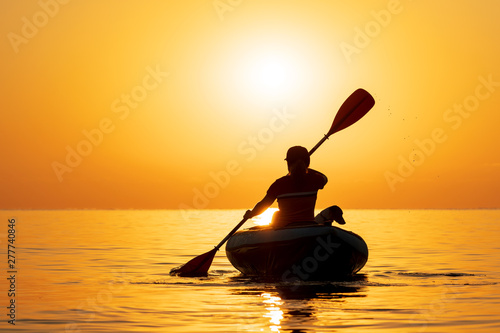 Red-haired young woman is rowing on an inflatable kayak by the sea with a dog Jack Russell Terrier on a background of pink sunrise in beautiful nature. Great disk of the rising sun. Sun rays. Sport