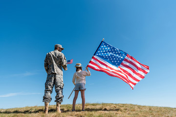 soldier in military uniform standing near kid with american flag against blue sky