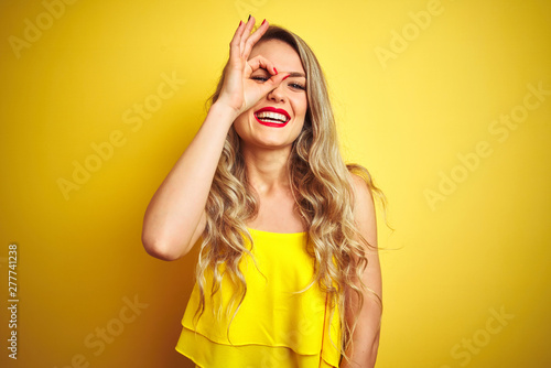 Young attactive woman wearing t-shirt standing over yellow isolated background doing ok gesture with hand smiling, eye looking through fingers with happy face.