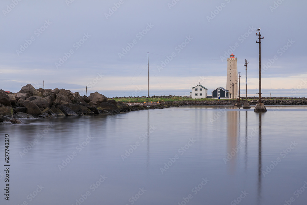 Blue sky with power pylons in the water to the lighthouse and uninhabited houses. Stone coastline of the island of Grotta near Reykjavik with clouds. Peninsula with bird sanctuary