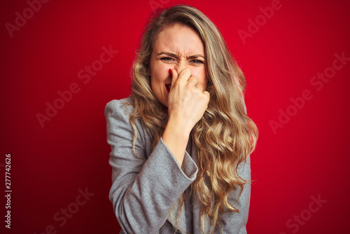 Young beautiful business woman wearing elegant jacket standing over red isolated background smelling something stinky and disgusting, intolerable smell, holding breath with fingers on nose. 