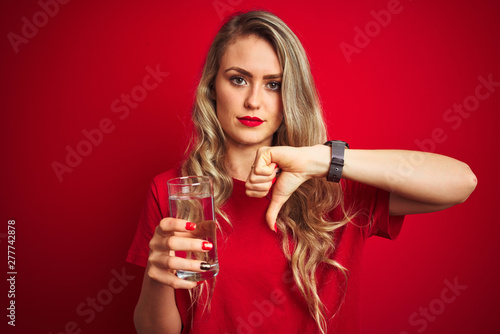 Young beautiful woman drinking a glass of water over red isolated background with angry face, negative sign showing dislike with thumbs down, rejection concept