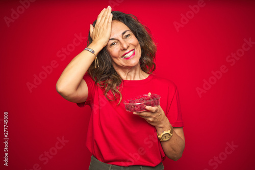 Middle age senior woman eating raspberries over red isolated background stressed with hand on head, shocked with shame and surprise face, angry and frustrated. Fear and upset for mistake.