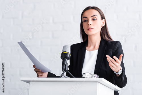 Fototapeta confident lecturer gesturing while speaking into microphone on podium tribune