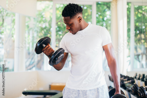 Young African American man standing and lifting a dumbbell with the rack at gym. Male weight training person doing a biceps curl in fitness center
